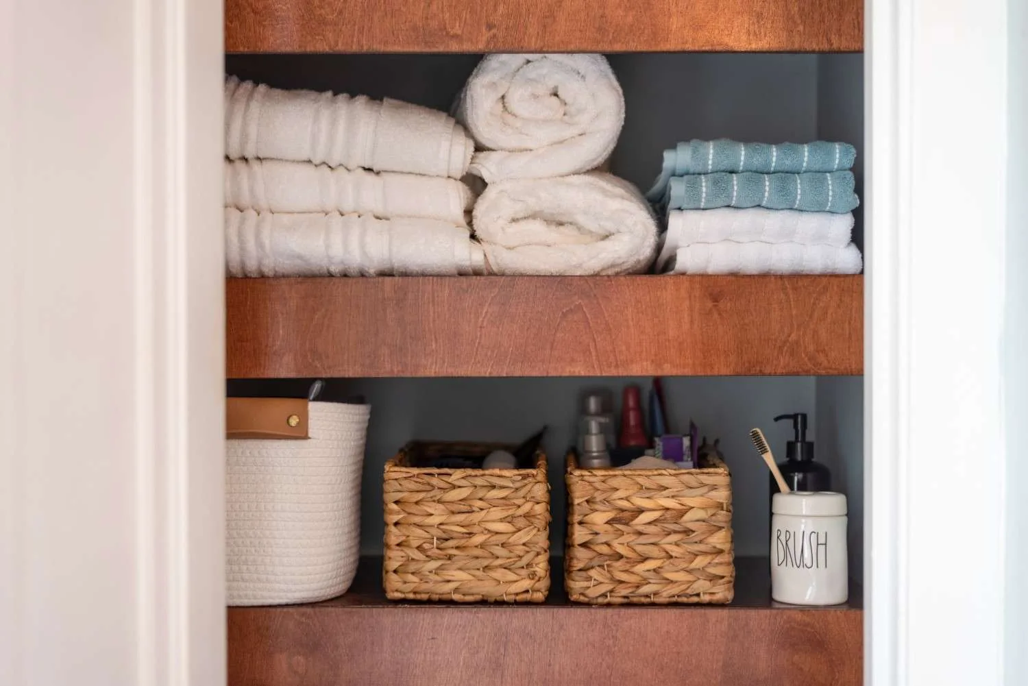 Neatly organized open wooden bathroom shelves featuring stacked white towels, rolled linens, and woven storage baskets containing toiletries.