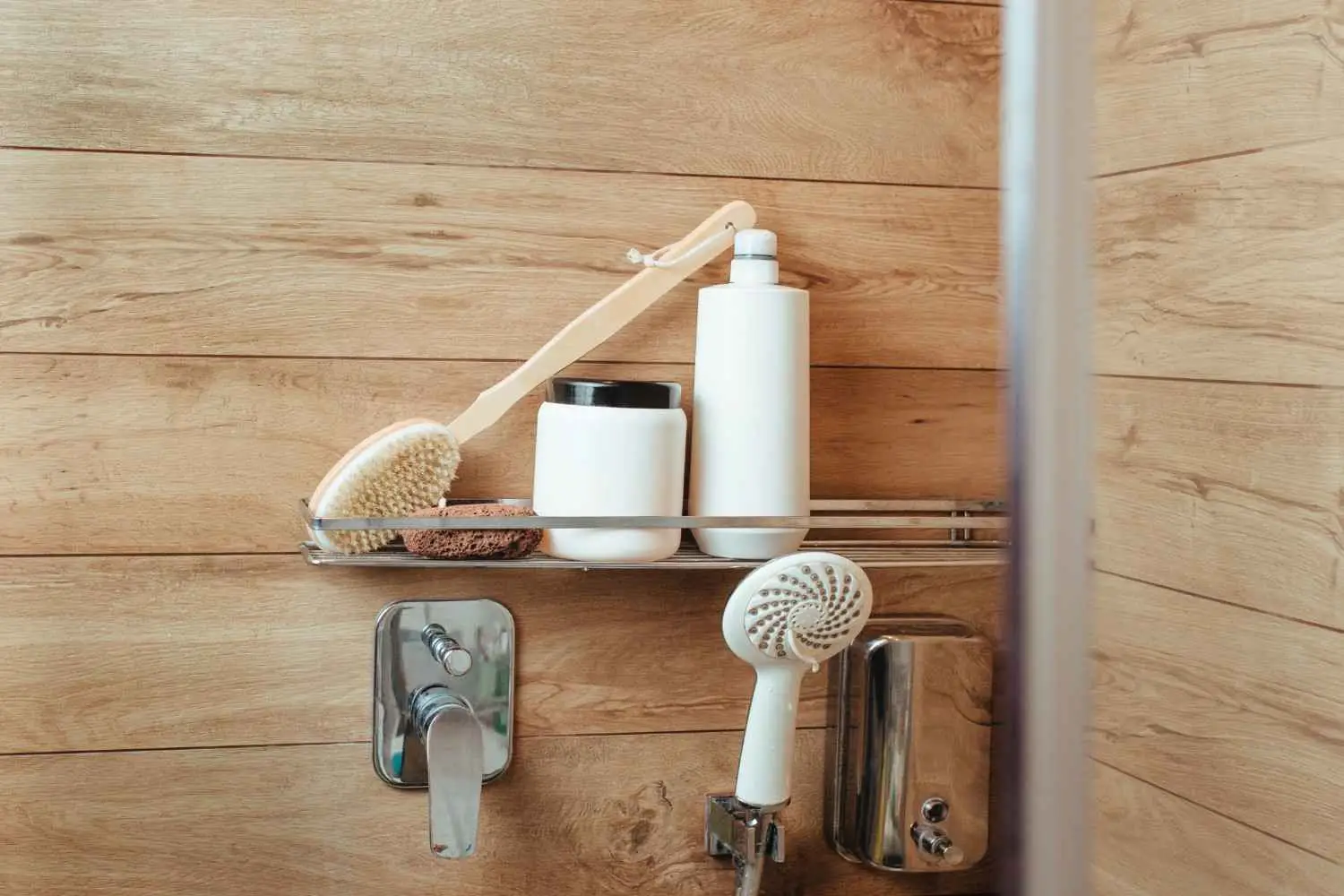 A sleek chrome shower caddy mounted on a wood-textured wall, holding a long-handled body brush, a pumice stone, and two white cosmetic bottles.