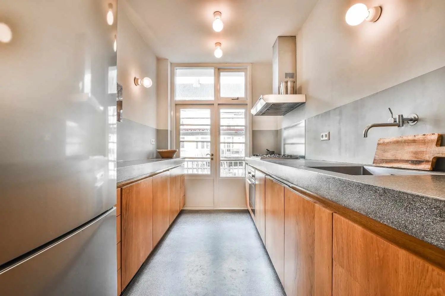 A narrow galley kitchen featuring warm wood cabinetry, gray concrete countertops, polished gray flooring, and minimalist globe pendant and wall lighting.