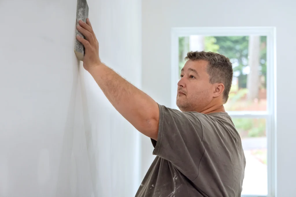 A professional worker sanding smooth drywall mud on a white wall near a window, illustrating the labor involved in determining a Drywall Repair Cost for a 2026 home renovation.