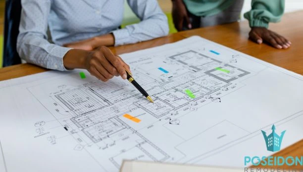 A close-up, midsection view of professional architects using a pen to point at detailed house floor plans on a wooden table, illustrating the first phase of Our Remodeling Process: Clear Steps, No Surprises.