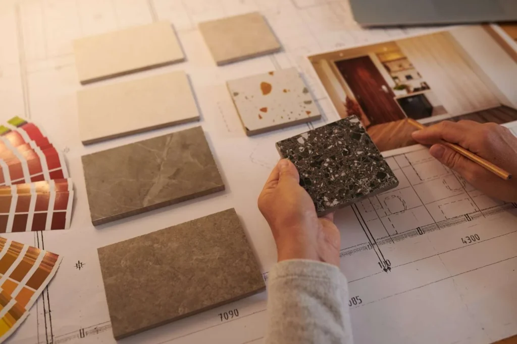 An interior designer designing a kitchen layout by comparing stone and terrazzo tile samples over a technical architectural floor plan.