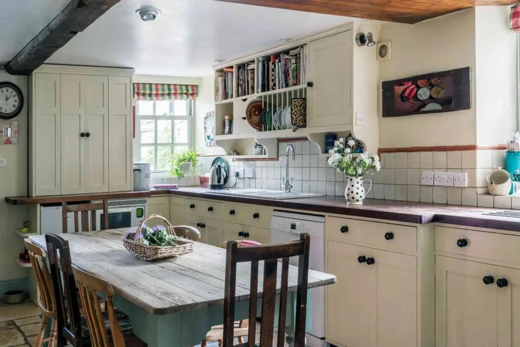 A rustic farmhouse kitchen with cream-colored shaker cabinets, a dark wood countertop, an exposed ceiling beam, and a weathered wood dining table with mismatched chairs.