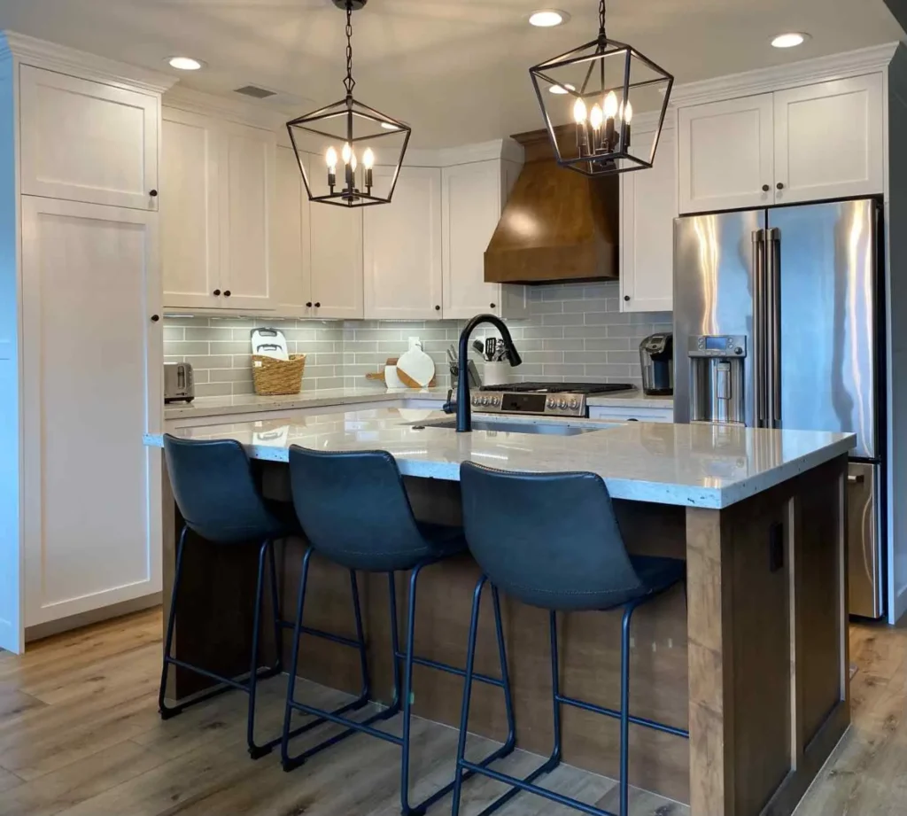 A small farmhouse kitchen featuring white shaker cabinets, a dark wood island with a white marble countertop, three black leather bar stools, and two geometric lantern pendant lights.