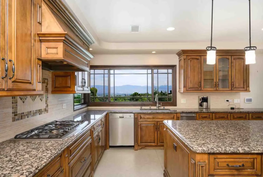 A spacious kitchen featuring ornate warm-toned wood cabinets, speckled granite countertops, a built-in gas stovetop, and a large window overlooking a mountain landscape.