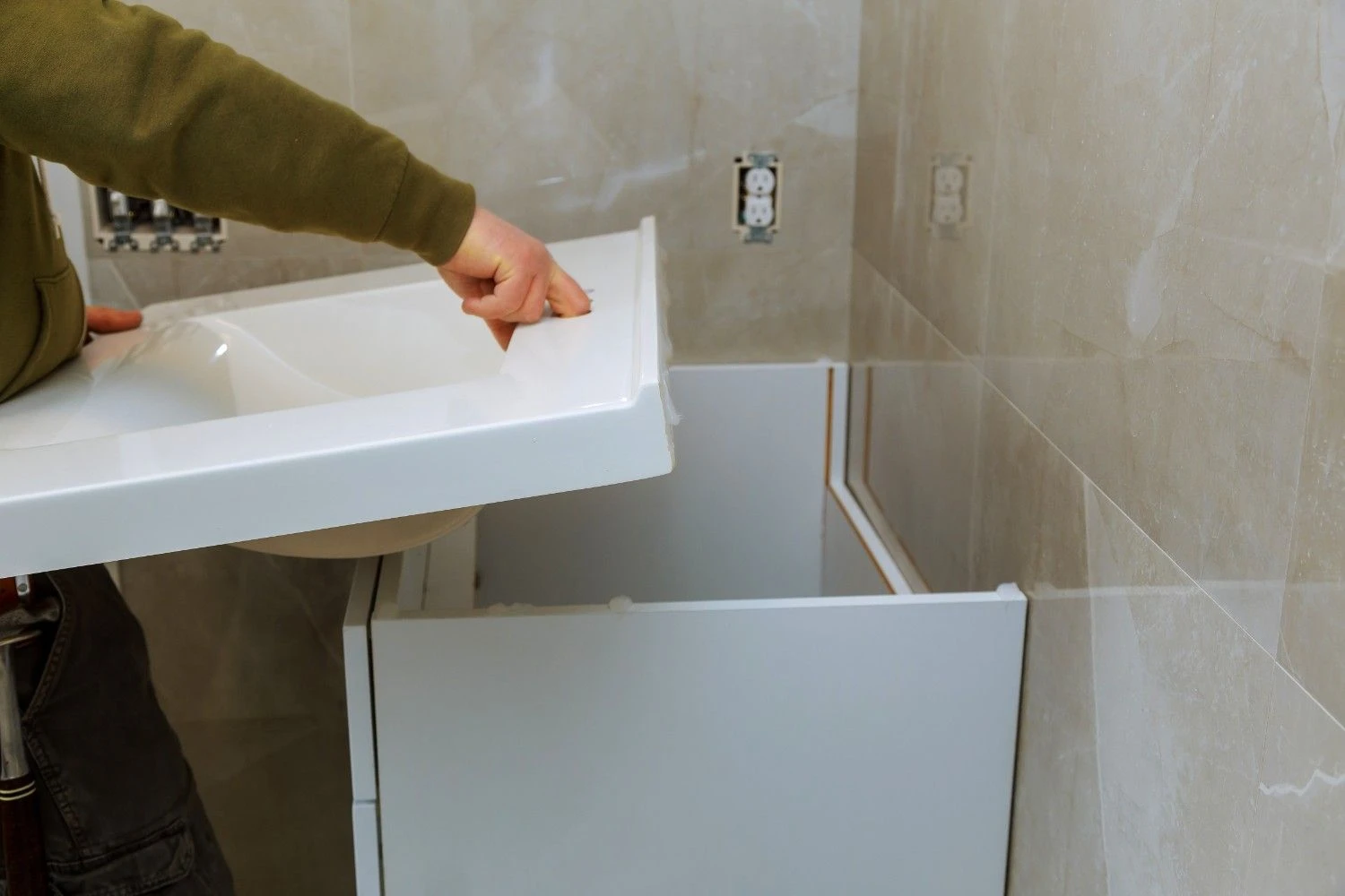 A contractor installing a white ceramic integrated sink and vanity top onto a white cabinet base against beige marble-patterned wall tiles.